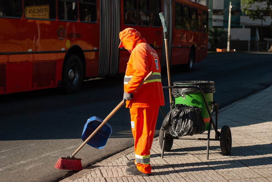A street scene featuring a municipal street cleaner dressed in bright orange high-visibility clothing, including a hooded jacket and reflective stripes. The worker is sweeping the pavement with a long