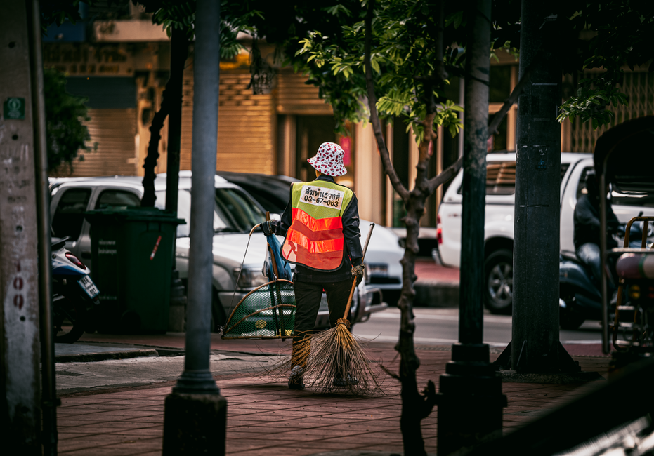 A street scene featuring a municipal street cleaner dressed in bright orange high-visibility clothing, including a hooded jacket and reflective stripes. The worker is sweeping the pavement with a long